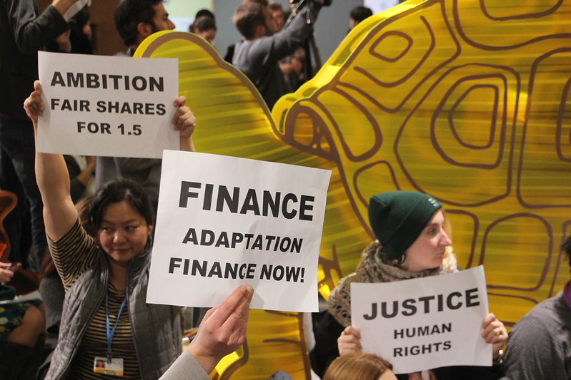 A small crowd of people holding up protest signs reading Ambition: Fair Shares for 1.5; Finance Adaptation now; Justice: Human Rights